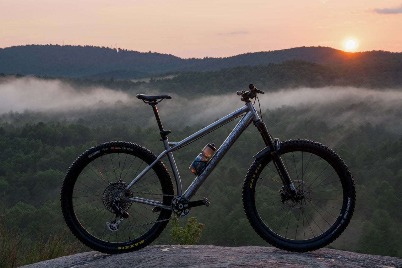 A full-suspension mountain bike with a silver frame and black tires is parked on a rock overlooking a misty forest valley at sunset. The sun is setting over distant mountains, with low-hanging fog filling the landscape below.